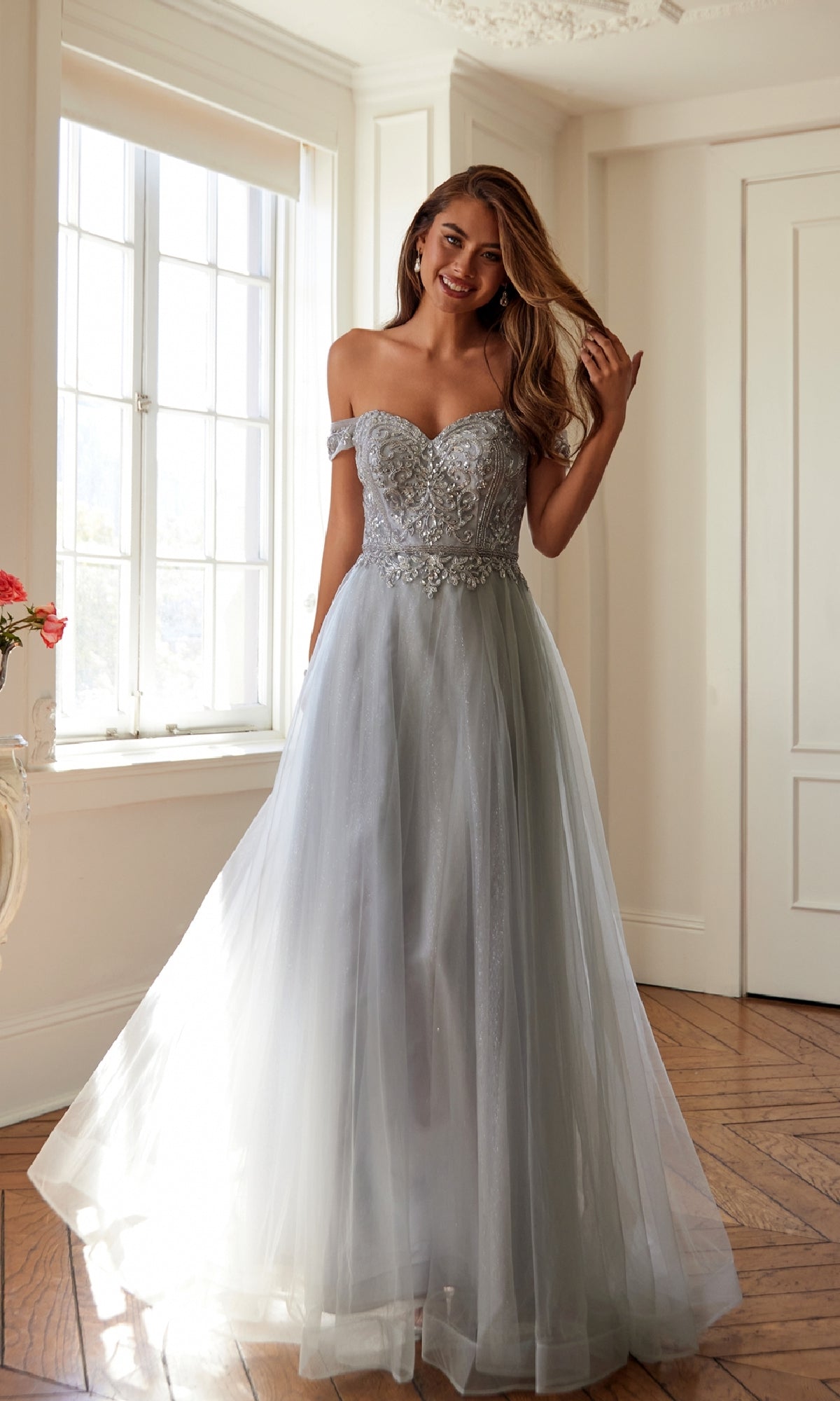 A woman stands indoors wearing the Juliet 287 Long Sweetheart Prom Ball Gown, featuring a light gray off-the-shoulder design with a beaded corset bodice and lace detailing. She smiles and holds her hair.